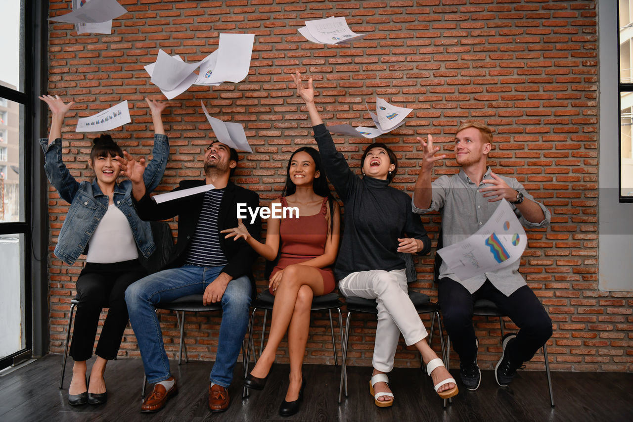 Cheerful coworkers throwing documents while sitting on chairs in office