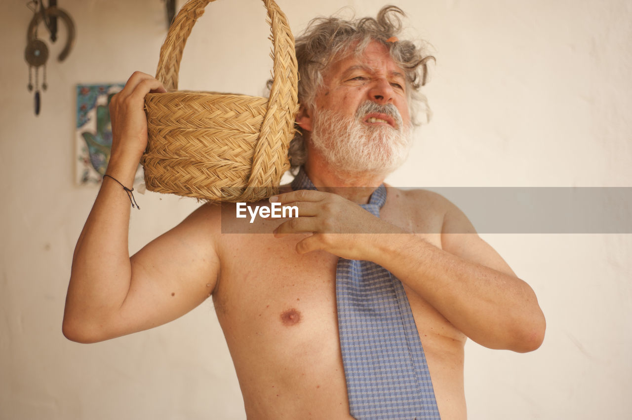 Shirtless man holding basket while standing against wall