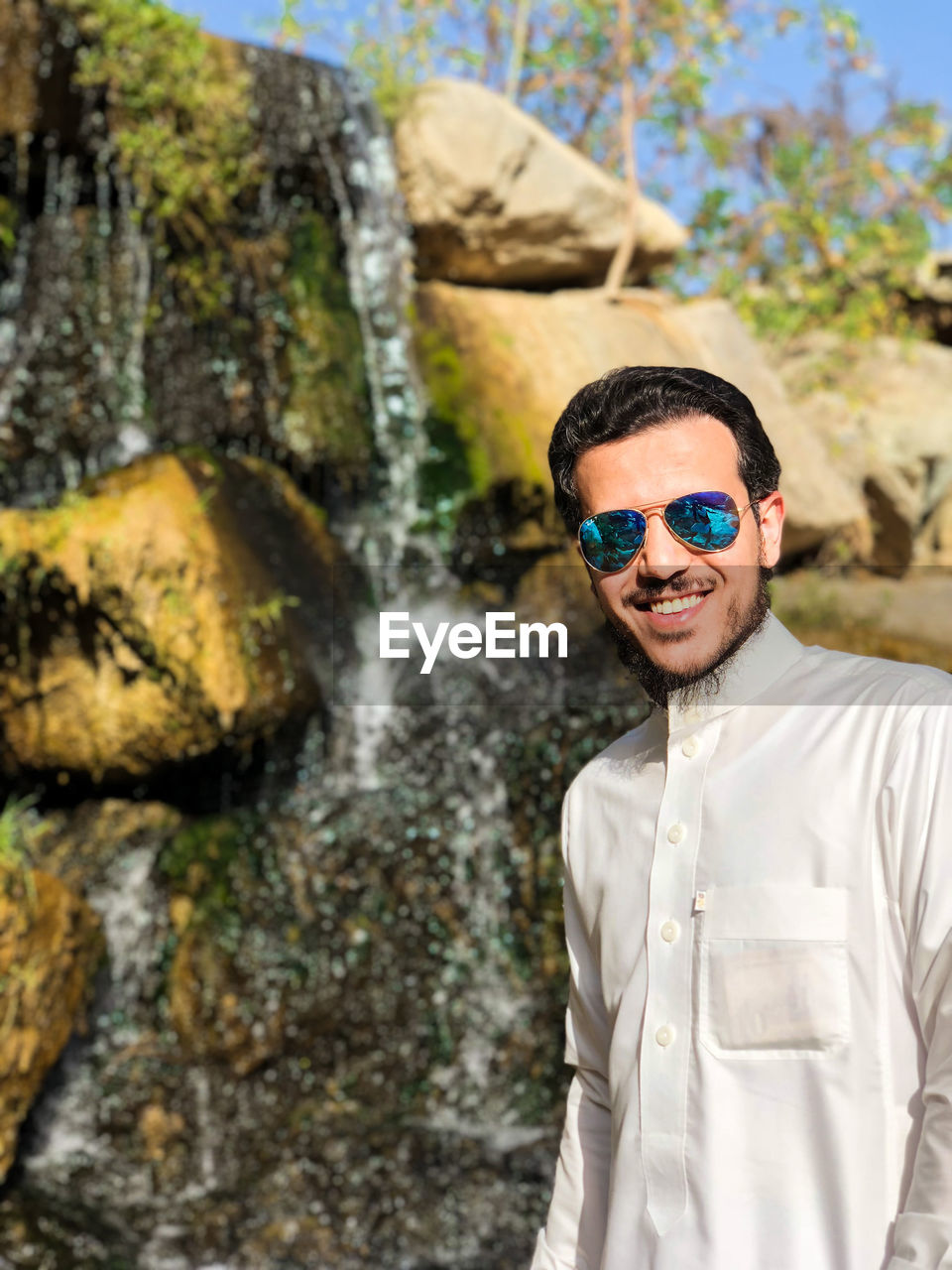 Portrait of young man wearing sunglasses standing against waterfall