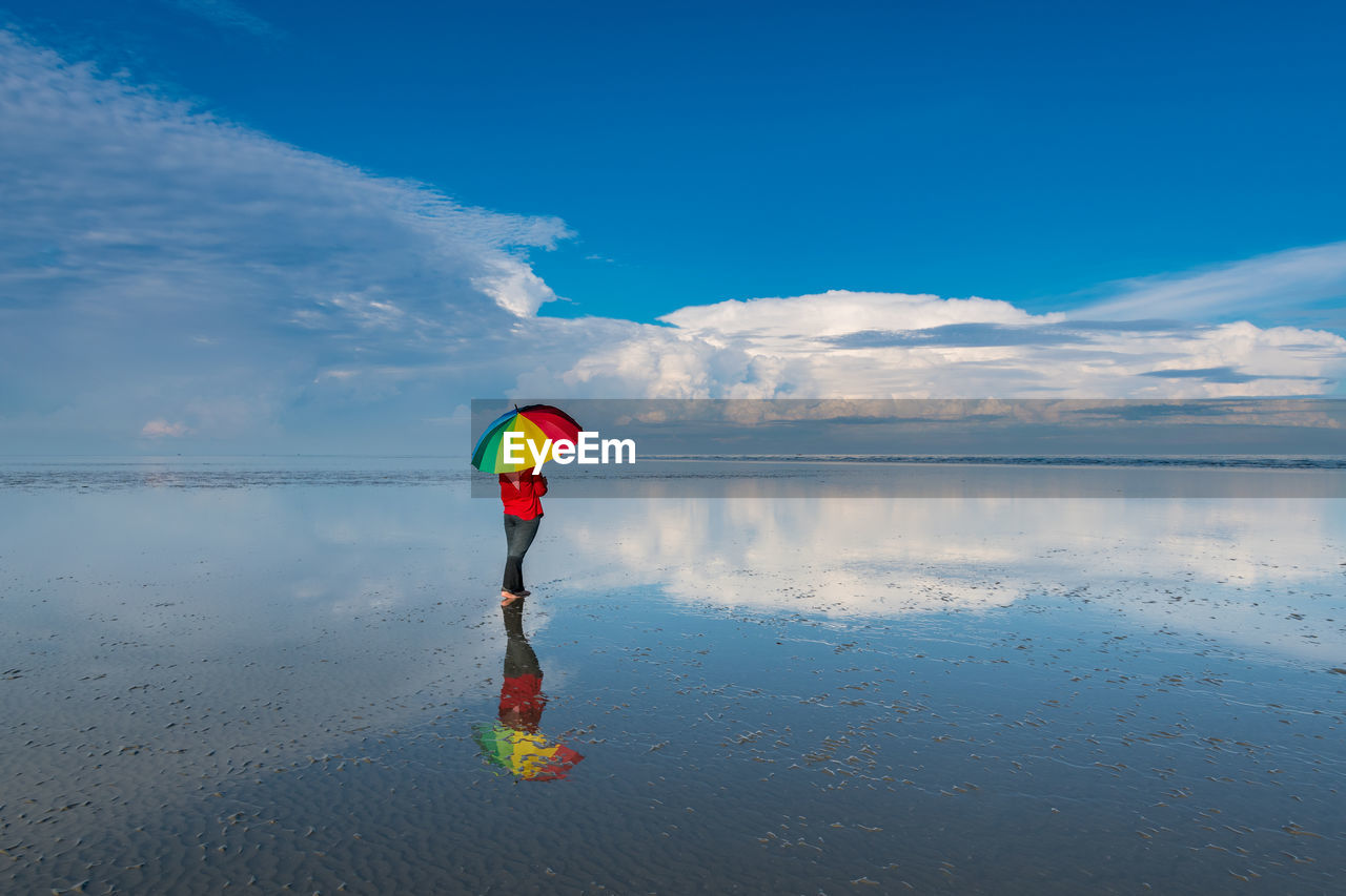 Rear view of woman with umbrella standing at beach against cloudy sky