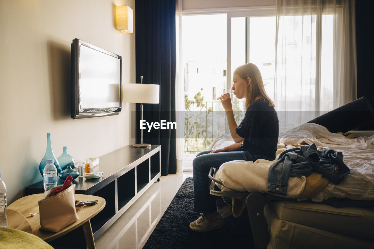 Side view of girl watching television while sitting on bed in hotel room