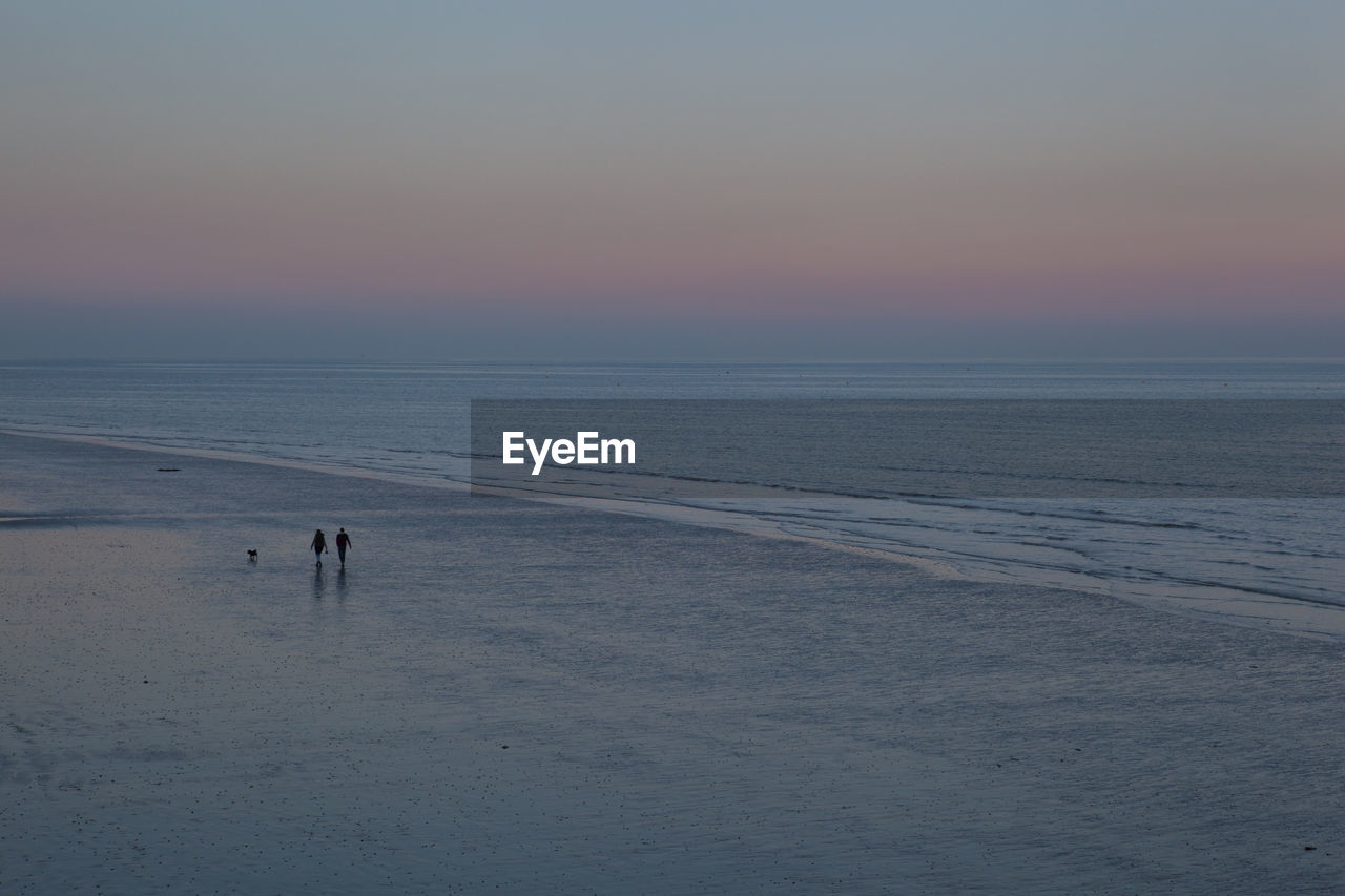 Distant view of people and dog at beach against sky during sunset