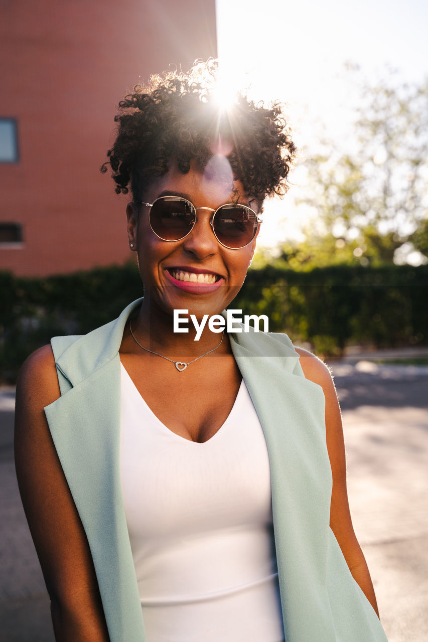 Joyful young african american female with afro hairdo and trendy sunglasses leaning elbow on railing and looking at camera while enjoying sunny summer day in park