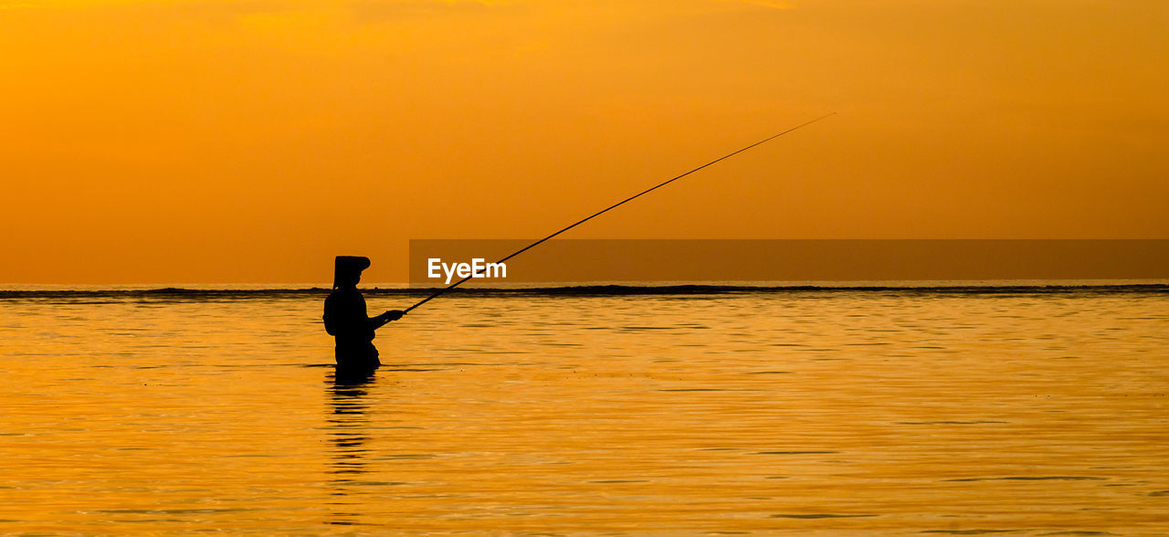 Silhouette fisherman fishing in sea against clear sky during sunset