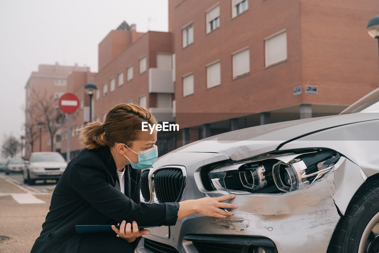 Side view of unrecognizable female insurance agent in formal outfit and protective mask writing protocol near damaged car parked on street after accident