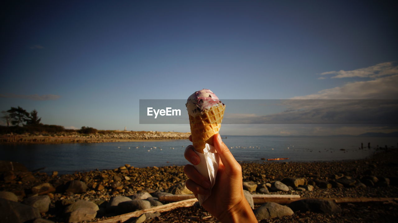 Woman holding ice-cream at beach against sky