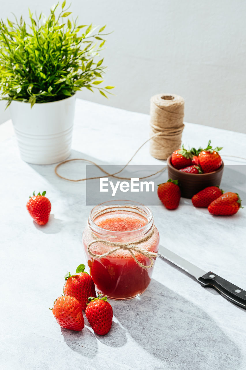 Composition with fresh homemade strawberry juice in glass jar wrapped with twine placed on marble surface with whole berries and knife