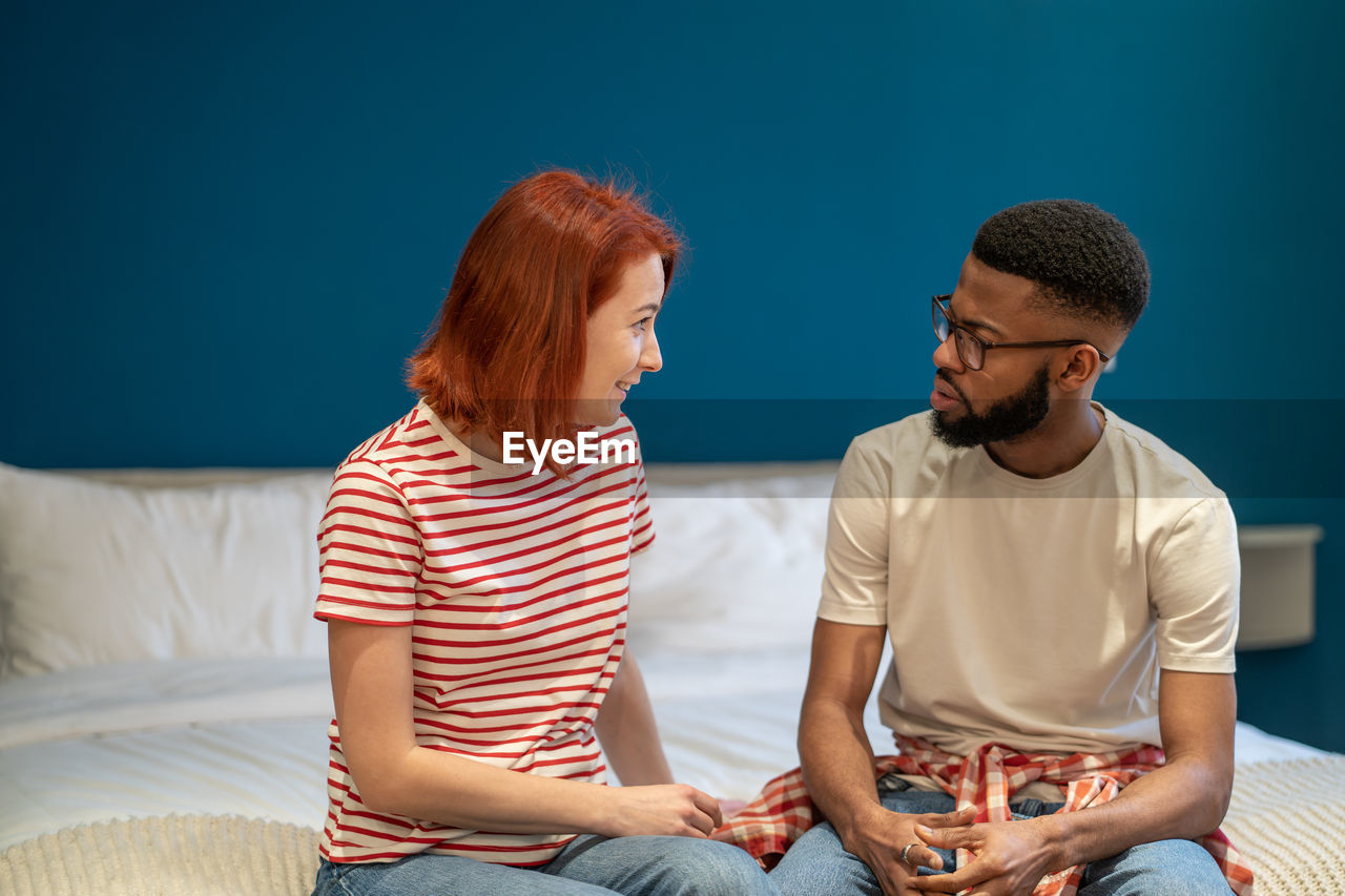 Young interracial couple husband and wife sit on bed at home discussing intimacy issues