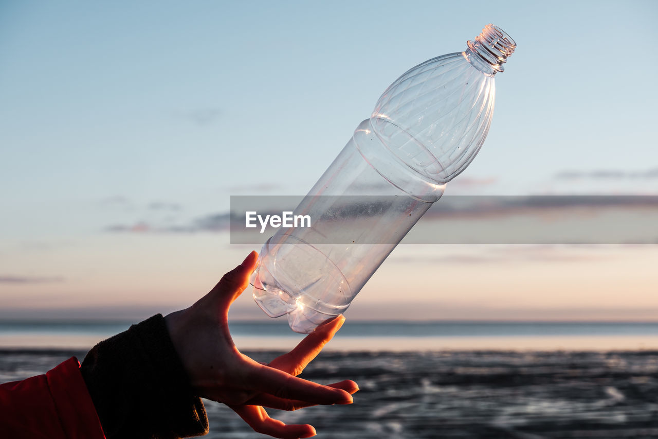 A human hand holds a plastic bottle thrown on the shore. 