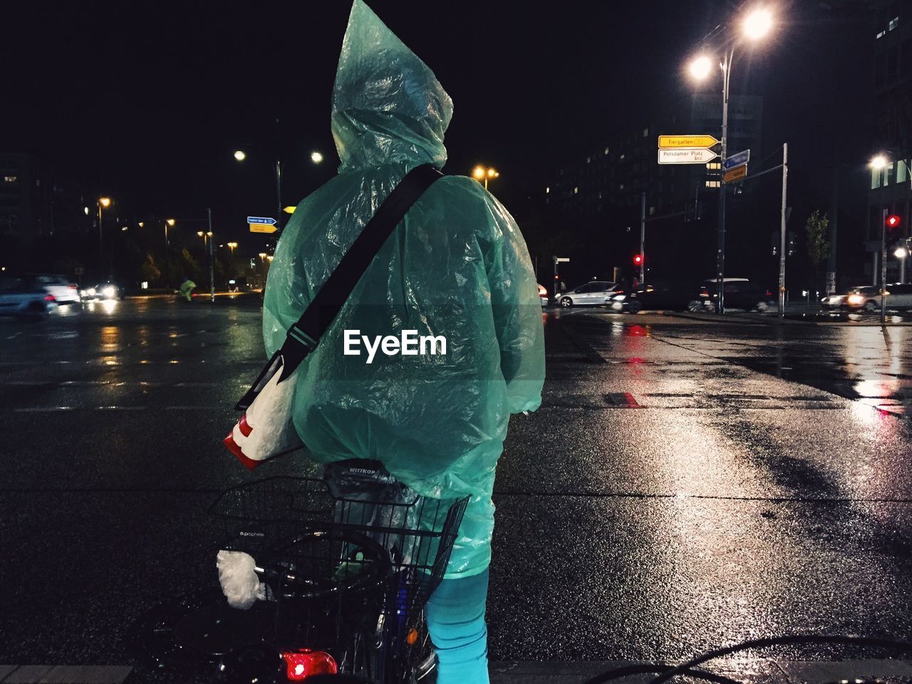 Rear view of woman in raincoat cycling on wet city street
