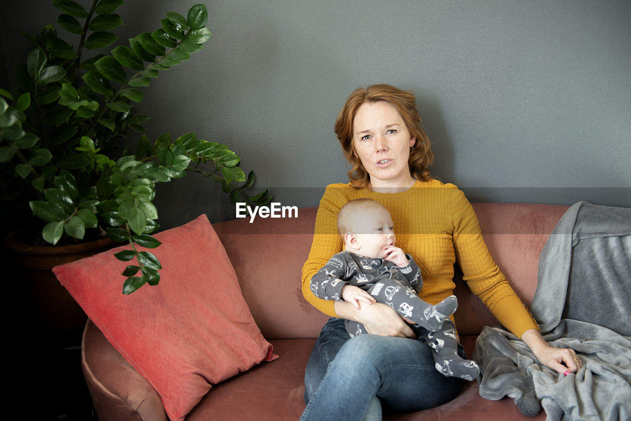 Serious grandmother holding baby grandson on sofa at home living room