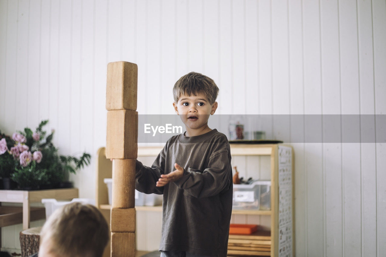 Boy looking away while standing near tower of toy blocks in classroom at kindergarten