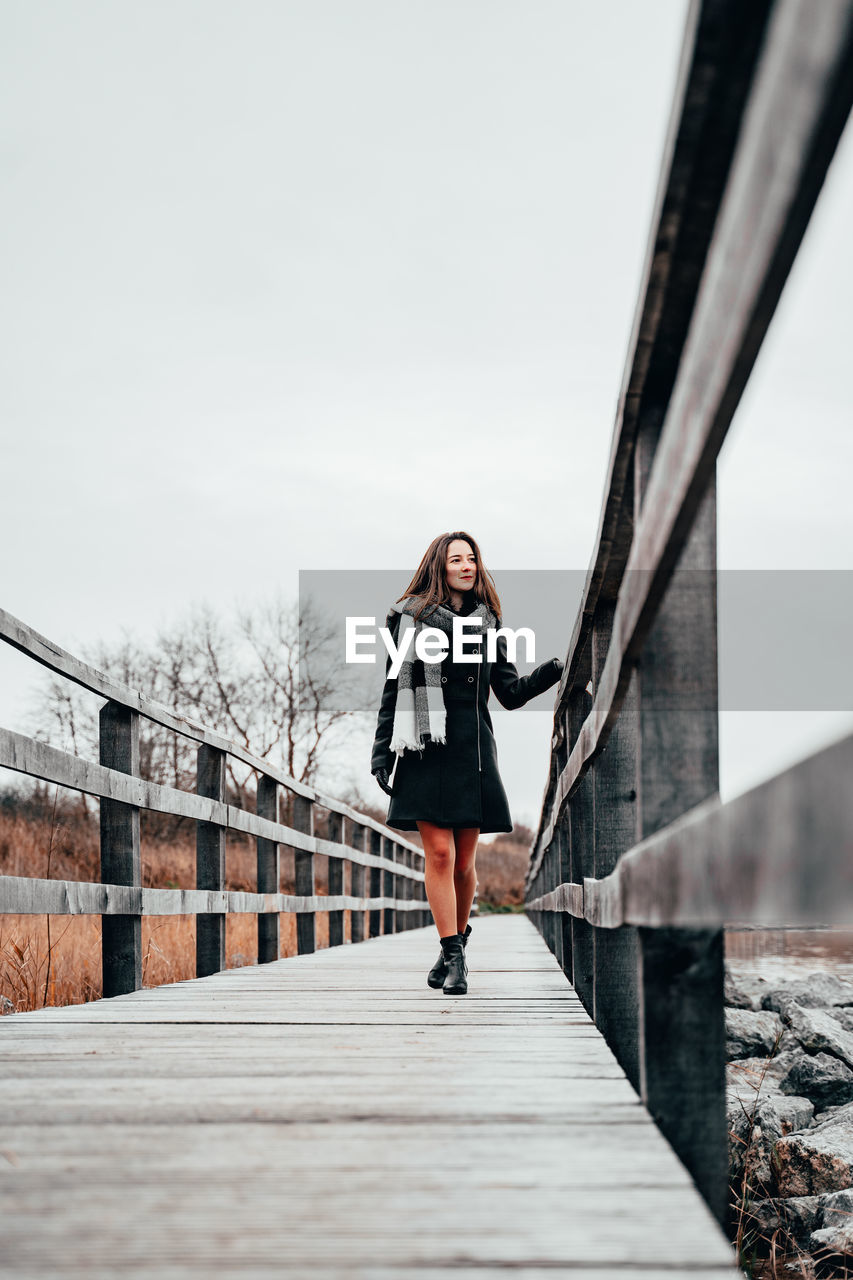 WOMAN STANDING ON FOOTBRIDGE AGAINST SKY