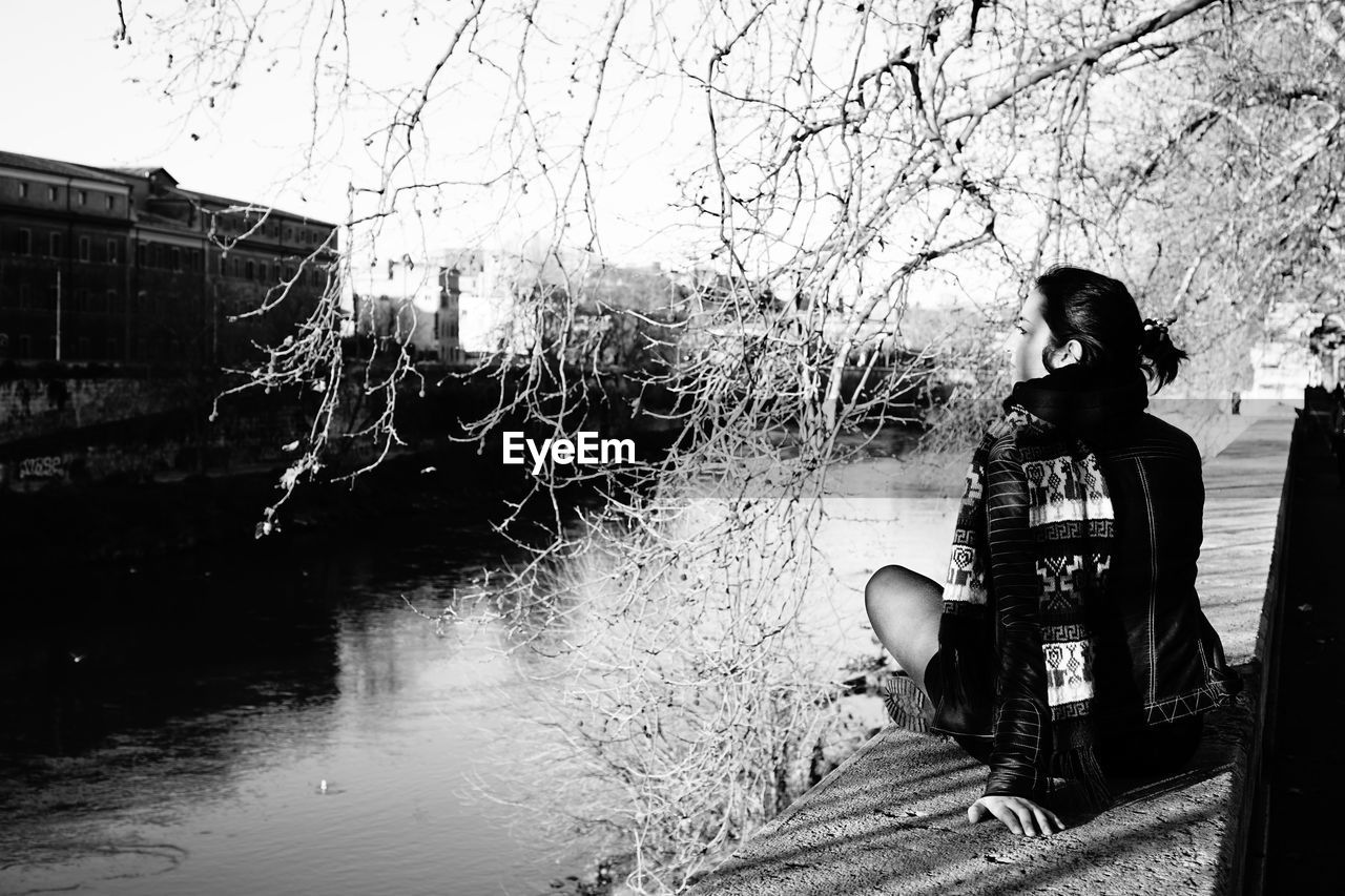 Rear view of woman sitting on retaining wall by river against sky