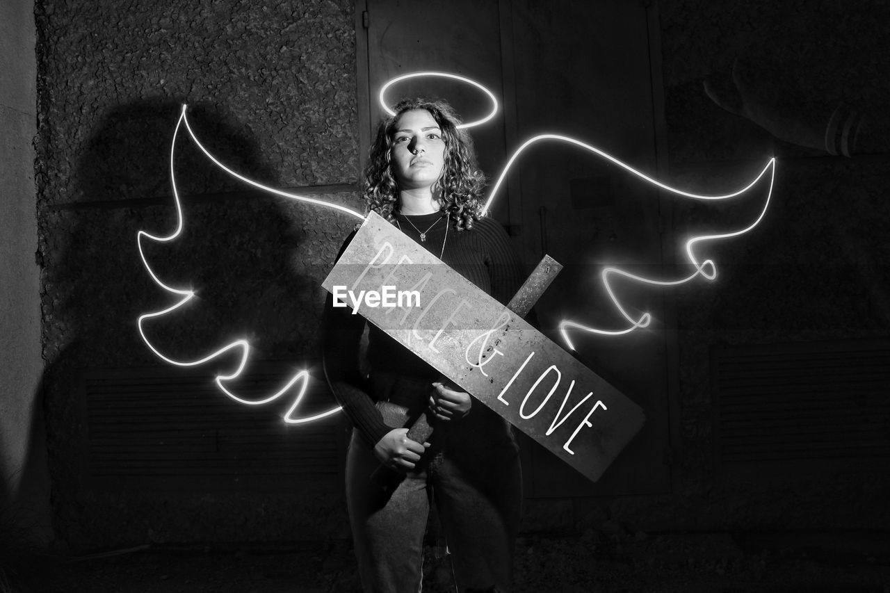 Digital composite image of woman holding information sign with wings and halo against wall