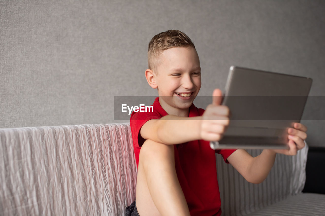 A happy boy in a red t-shirt is sitting on the couch with a tablet and laughing