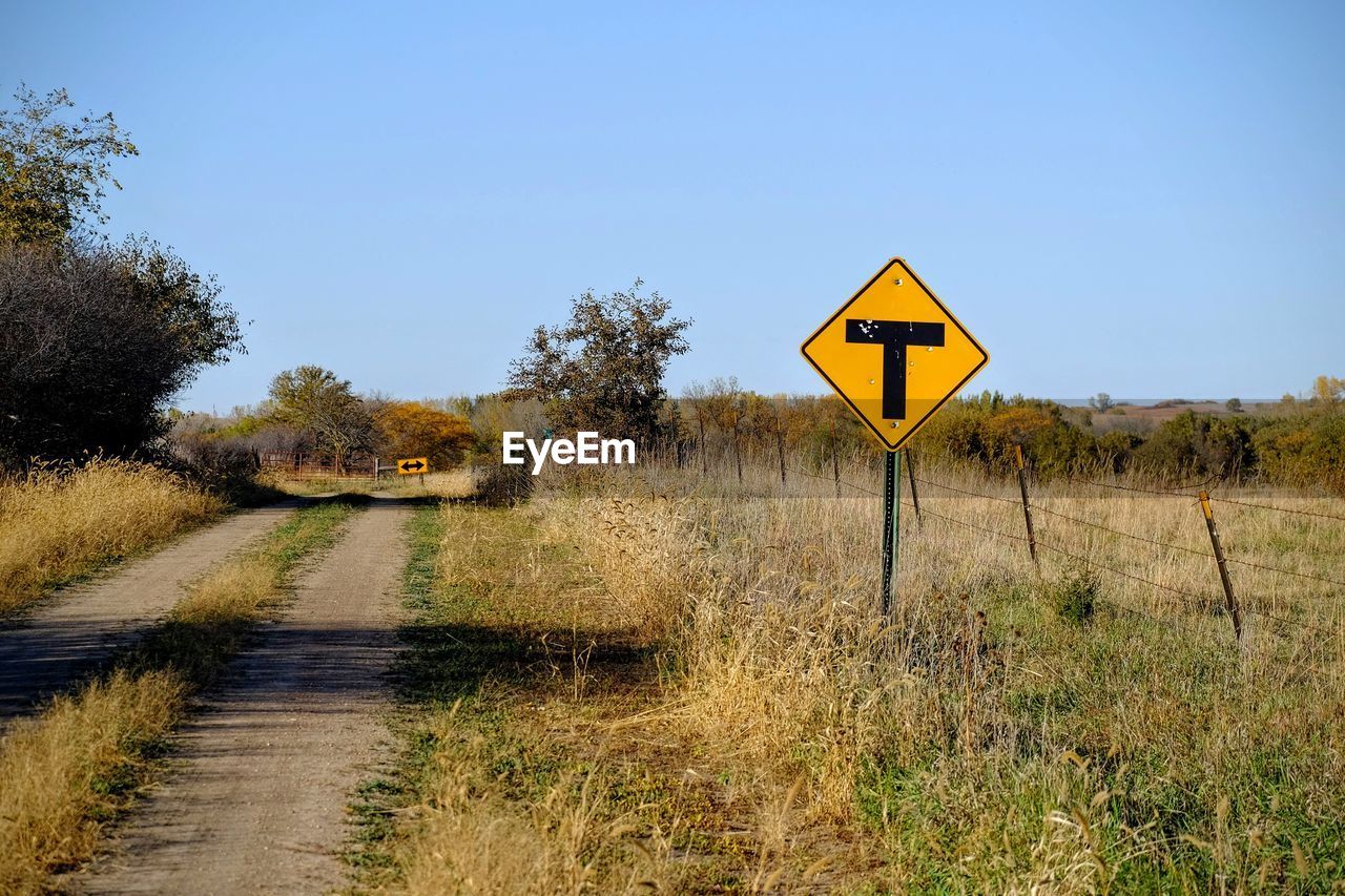 T intersection sign on grassy field by dirt road against blue sky