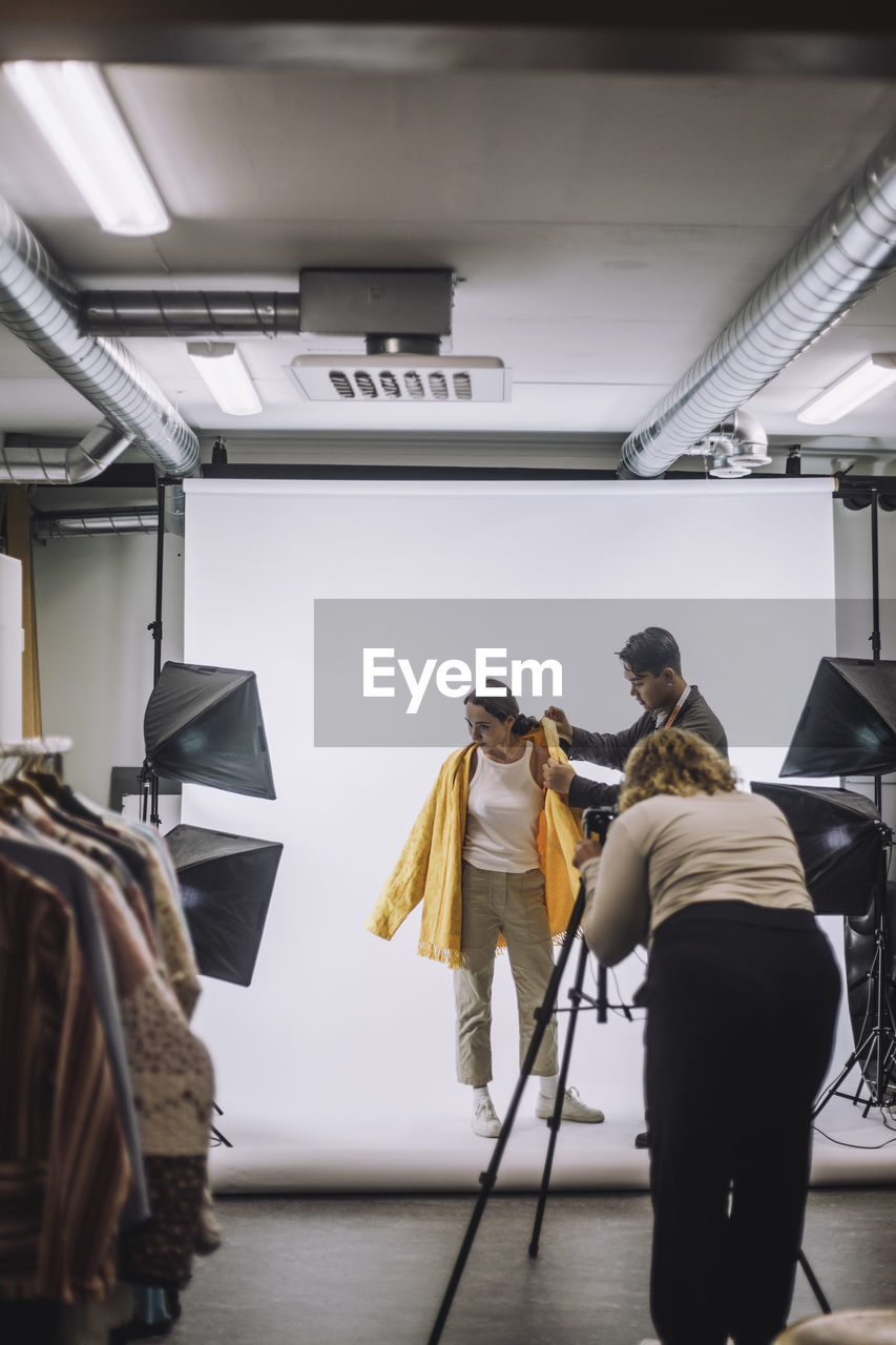 Photographer photographing designer helping model wearing jacket against white backdrop in studio