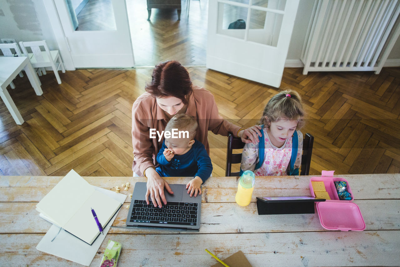 High angle view of mother working on laptop while sitting with cute son by daughter using digital tablet at table