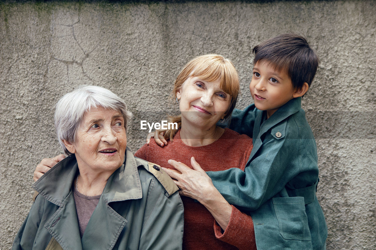 Three generations grandmother great grandmother grandson are standing on the street against a gray 