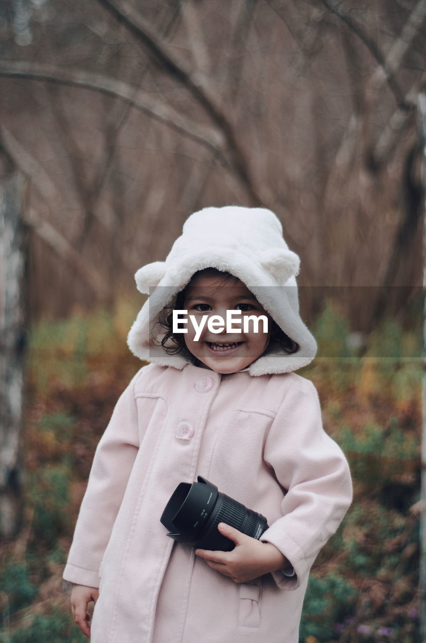 Cute girl in warm clothing holding camera lens while standing on field