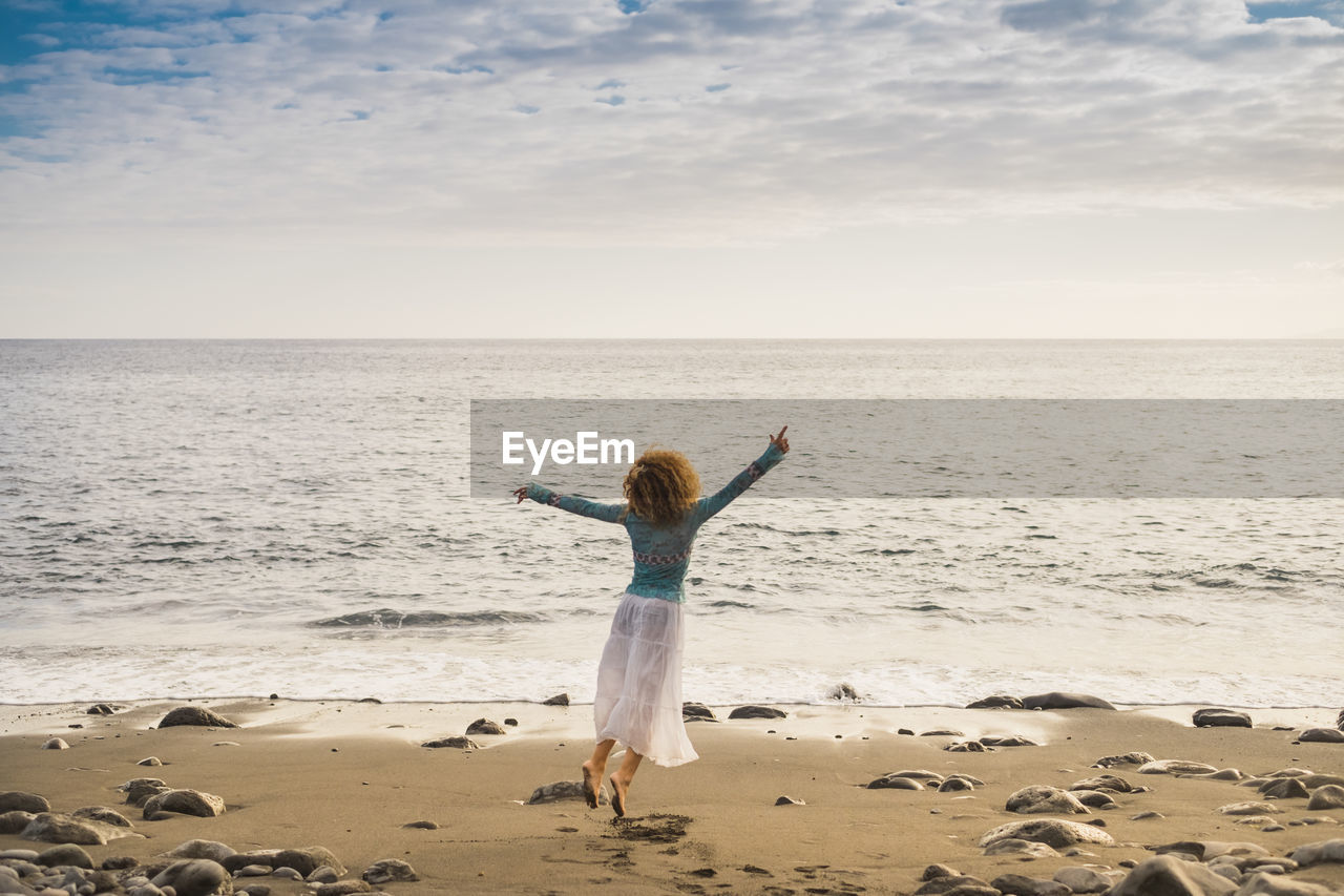 Rear view of woman dancing at beach against cloudy sky