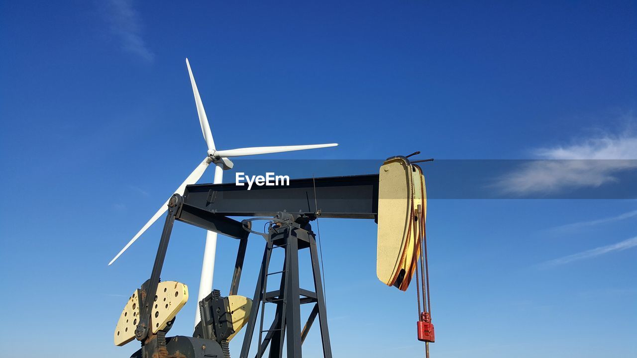 Low angle view of oil drill and wind turbine against blue sky