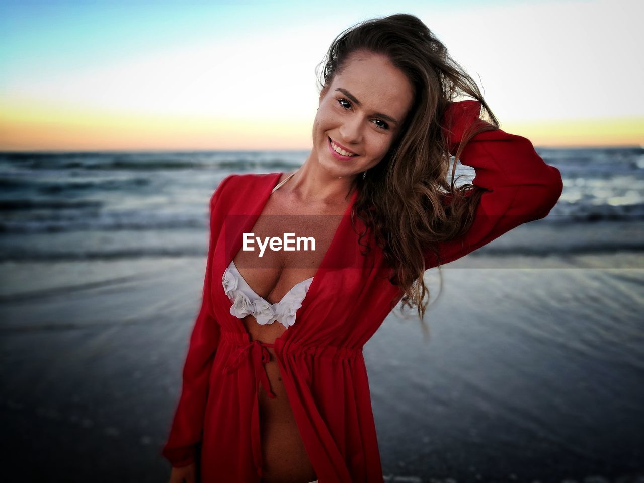 Portrait of beautiful woman standing with hand in hair on shore at beach against sky during sunset