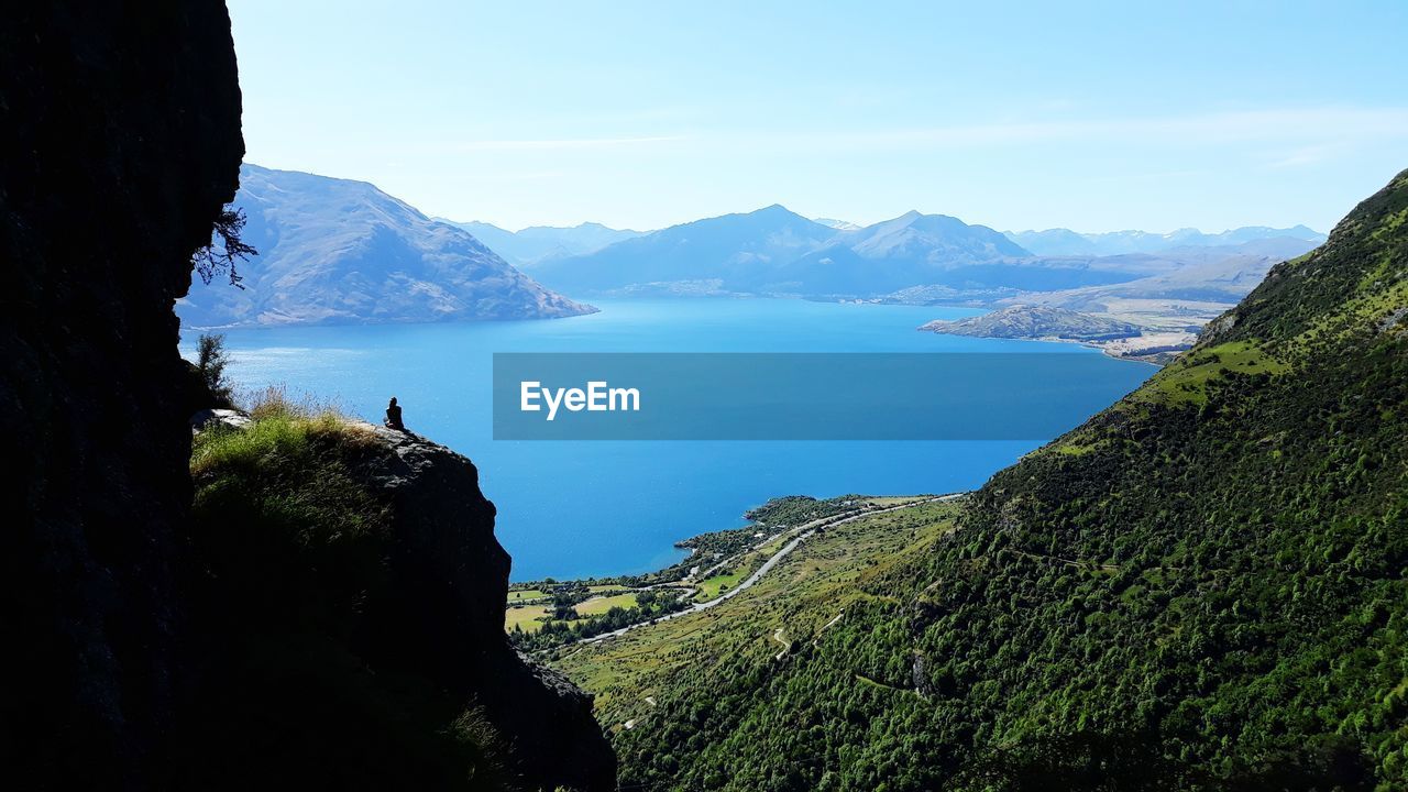 Mid distant view of woman sitting on cliff against lake and sky