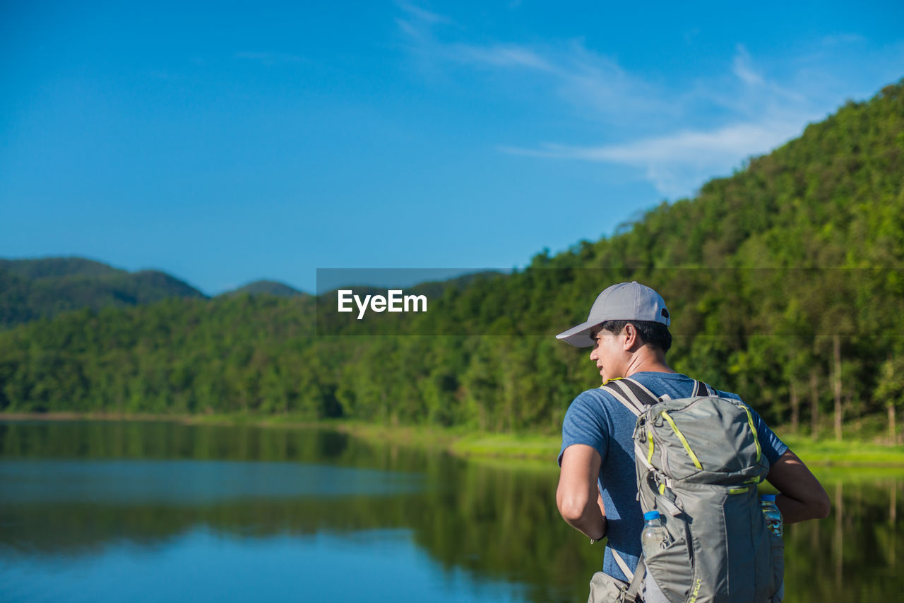 Rear view of man with backpack standing by lake against trees