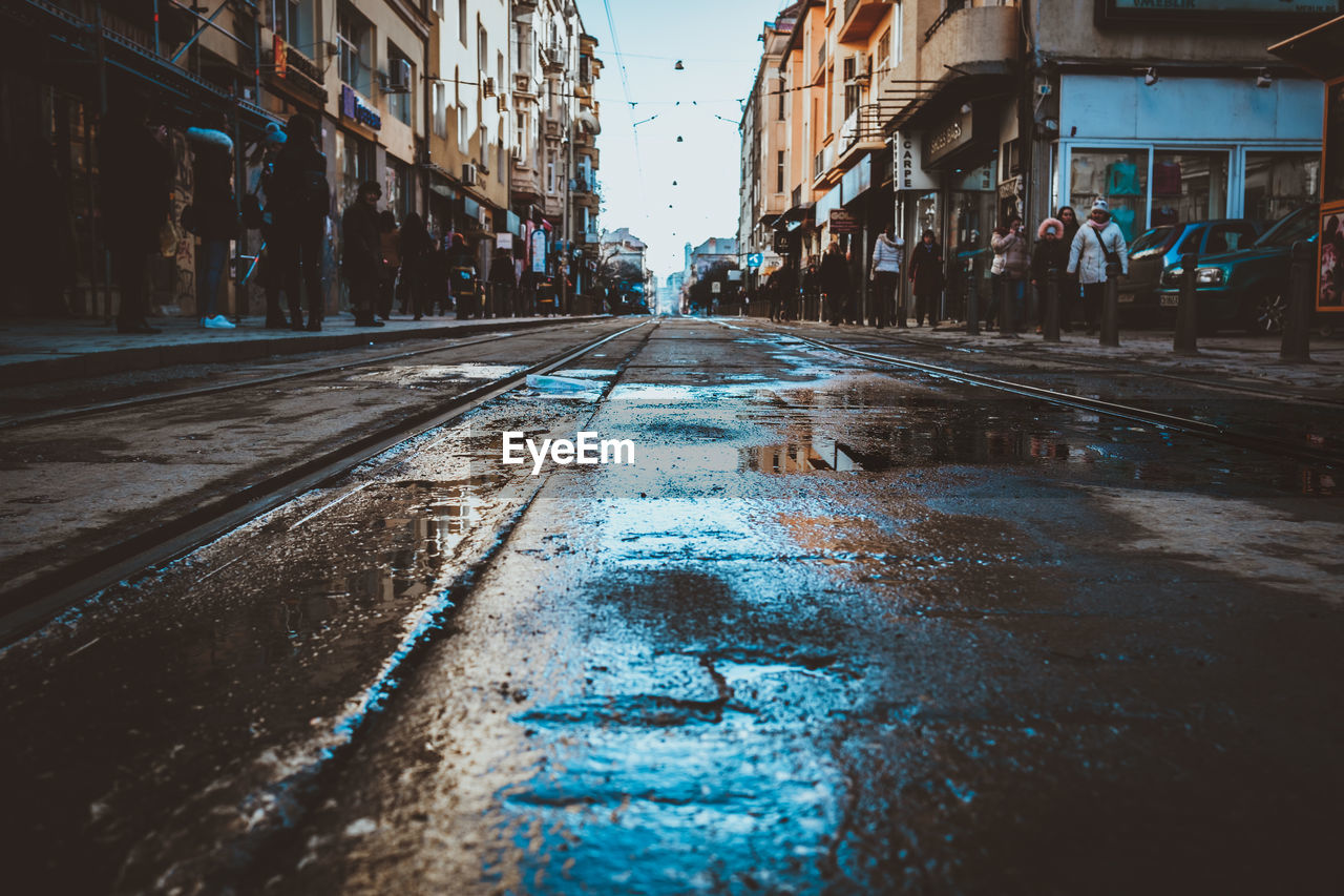 Wet street amidst people walking on footpath in city