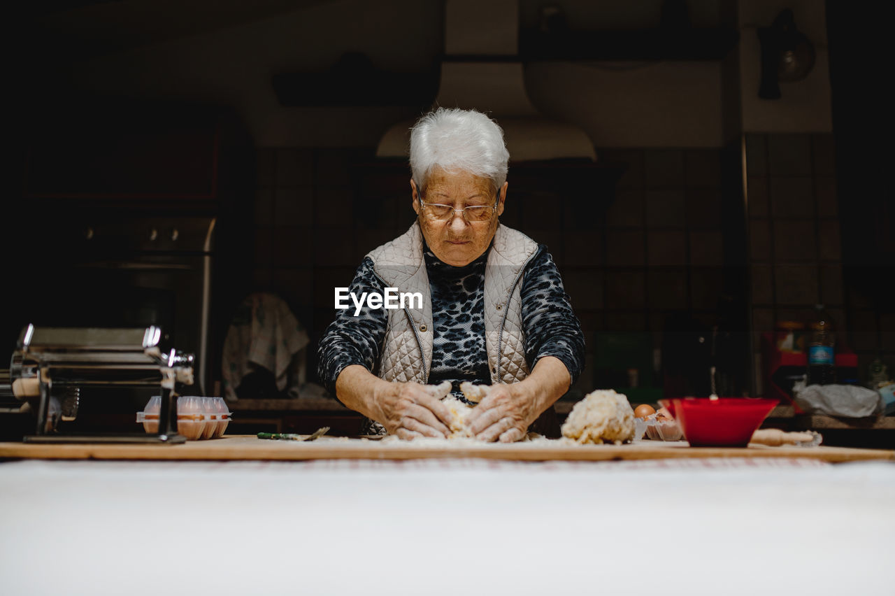 Senior female standing at table in kitchen and kneading dough while preparing ingredients for cooking appetizing tortellini