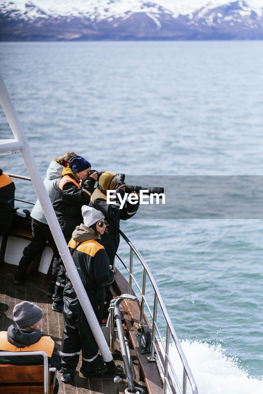 Side view of male and female travelers in protective clothes photographing and admiring seascape during cruise in modern boat on sunny day in iceland