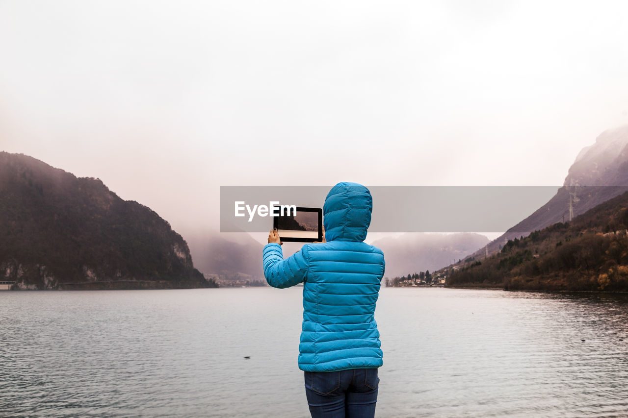 Rear view of woman photographing with digital tablet by lake during foggy weather