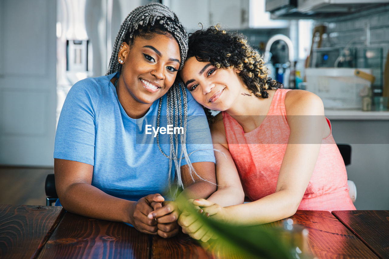 Portrait of confident happy lesbians sitting at table in kitchen