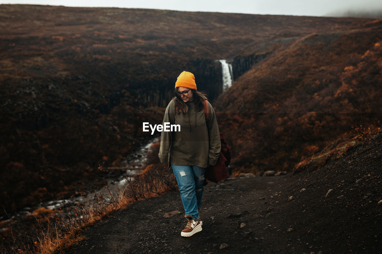 Young tourist in eyeglasses and hat with piercing looking down on hill near waterfall and mountain river