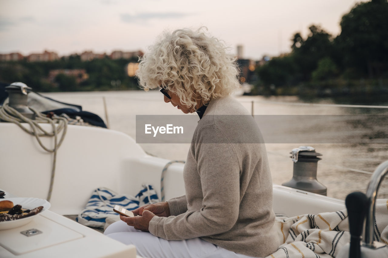 Senior woman text messaging on smart phone while sitting in boat during sunset