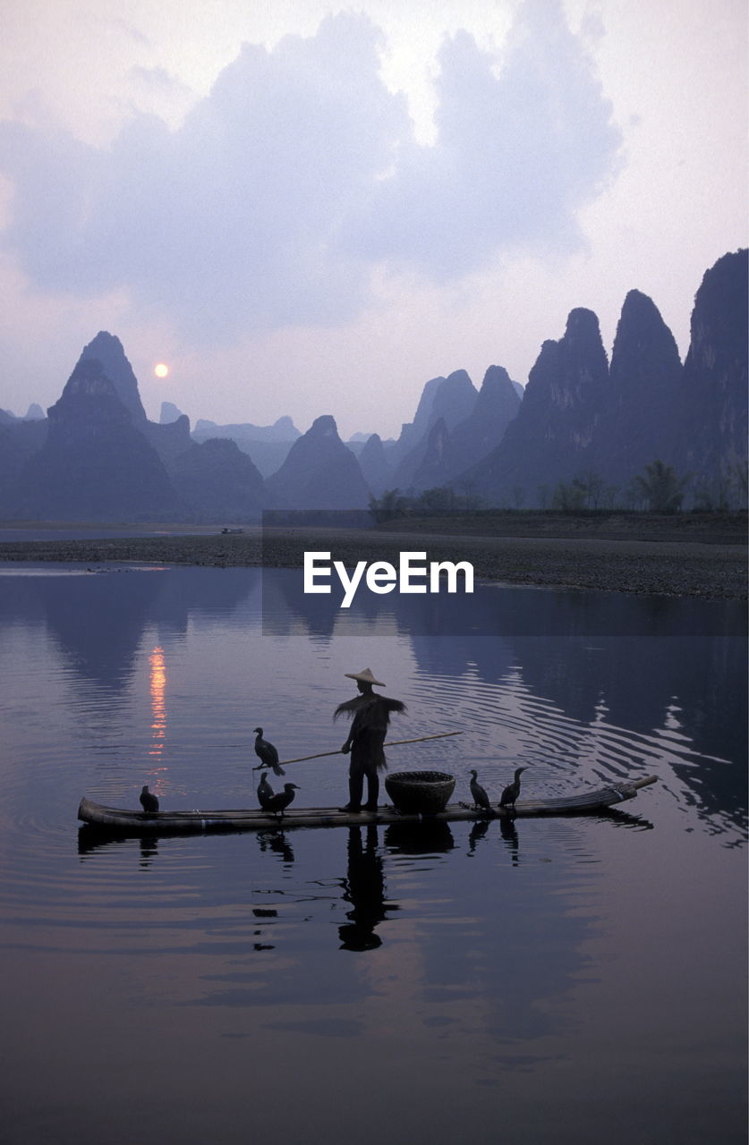 Man sailing boat with birds in river against mountains