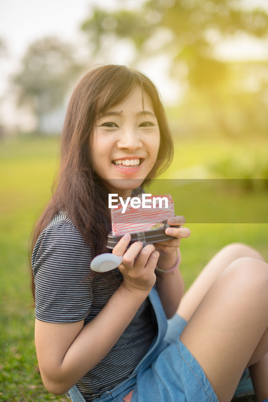 Portrait of smiling young woman holding pastry while sitting on field at park