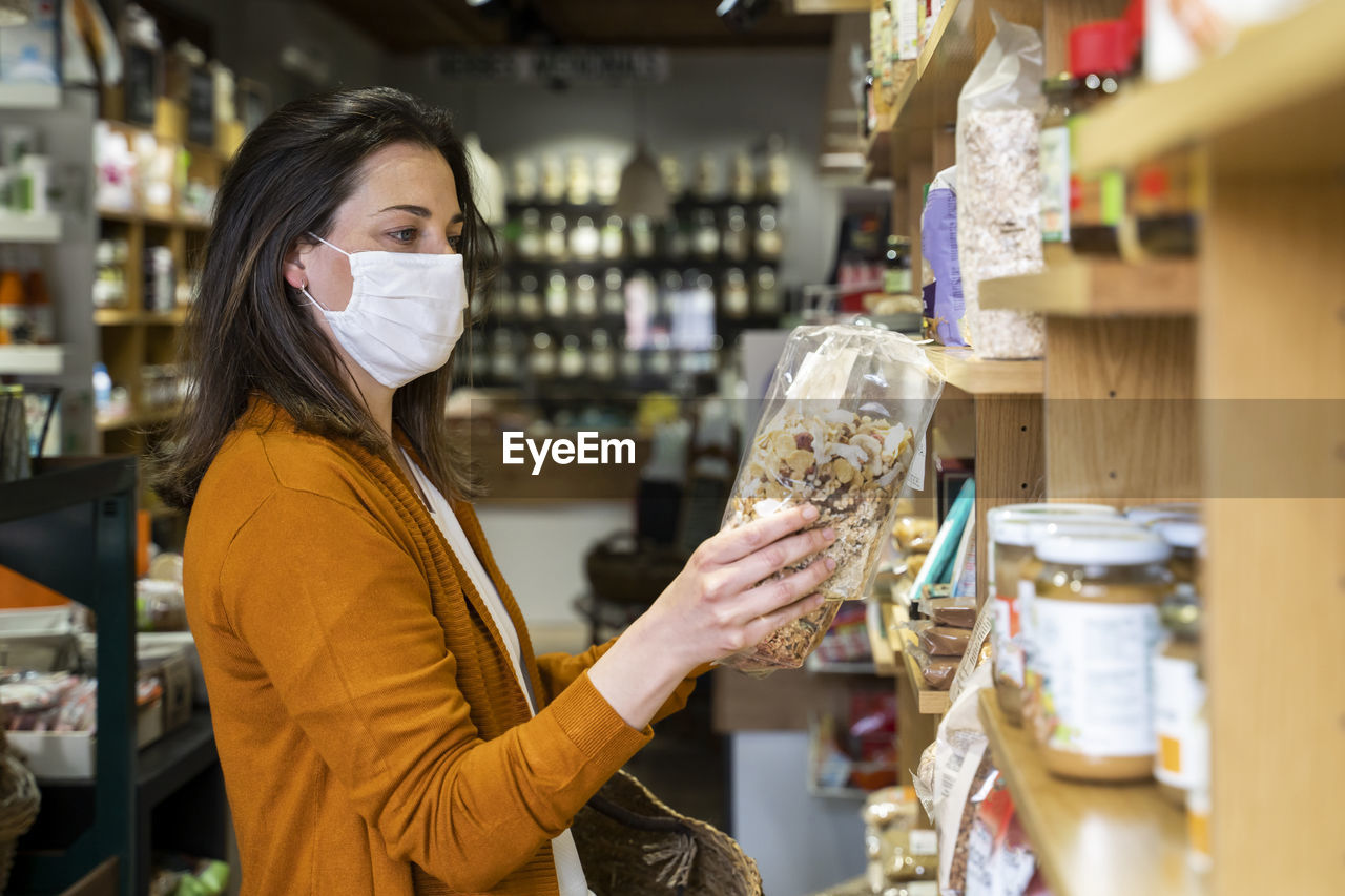 Young woman in protective face mask checking food package while shopping at grocery store