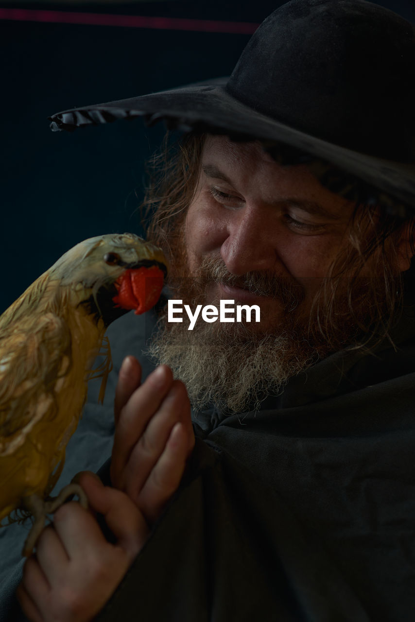 bird, one person, beard, adult, facial hair, portrait, men, animal, clothing, hat, animal themes, person, headshot, black background, mature adult, human face, one animal, emotion, indoors, dark, holding, animal wildlife, looking, close-up, falcon, studio shot