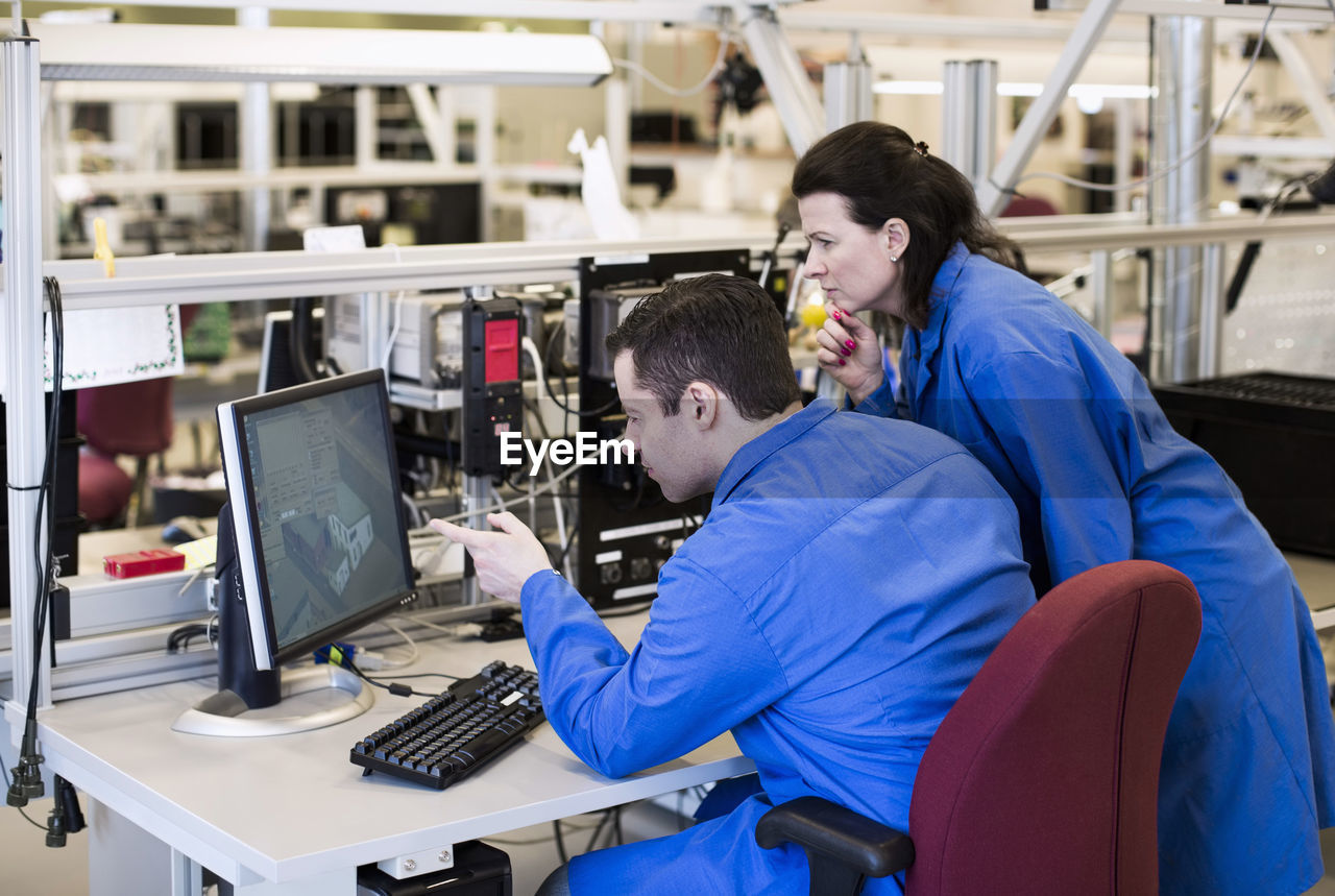 Male electrician pointing at computer monitor while discussing with colleague in industry