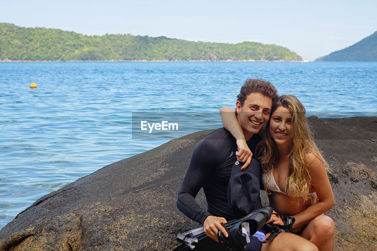 Portrait of smiling couple sitting on rock by sea