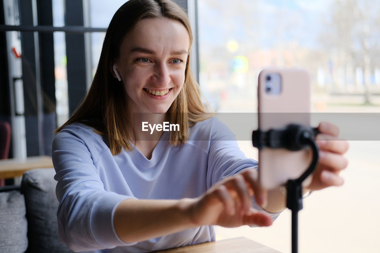 Young beautiful woman blogger making a video for her blog using phone camera in a cafe.