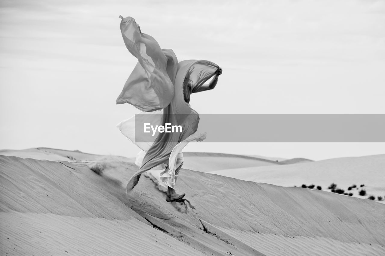 Woman with fabric jumping over sand at beach against sky