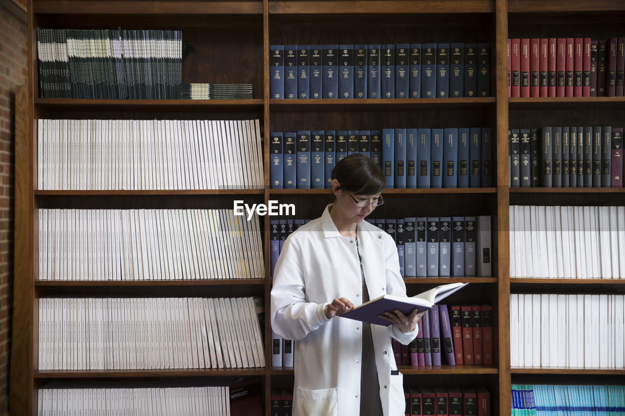 Female scientist reading book by shelves at laboratory