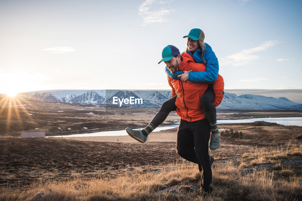 Man carrying woman on piggyback in field with mountains in background