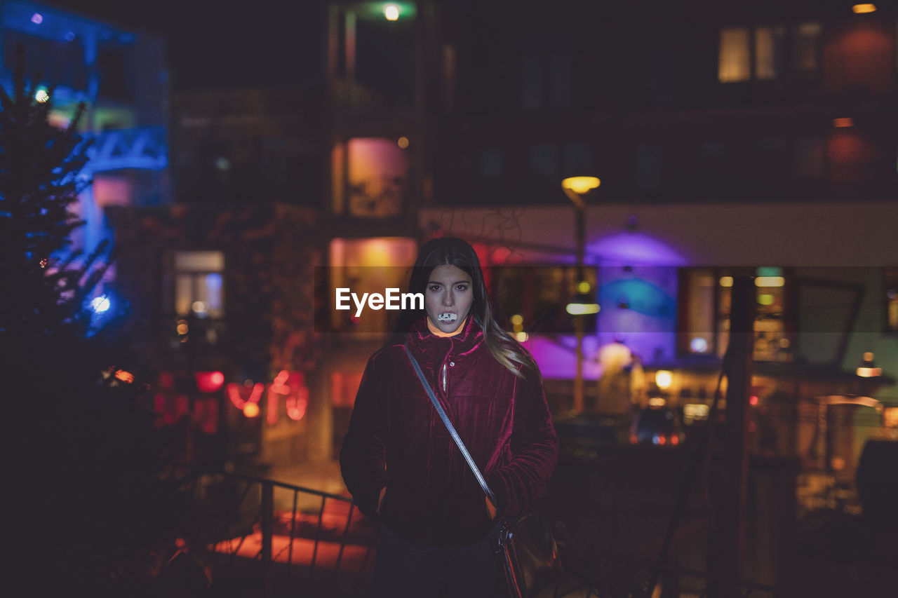 PORTRAIT OF WOMAN STANDING IN ILLUMINATED CITY