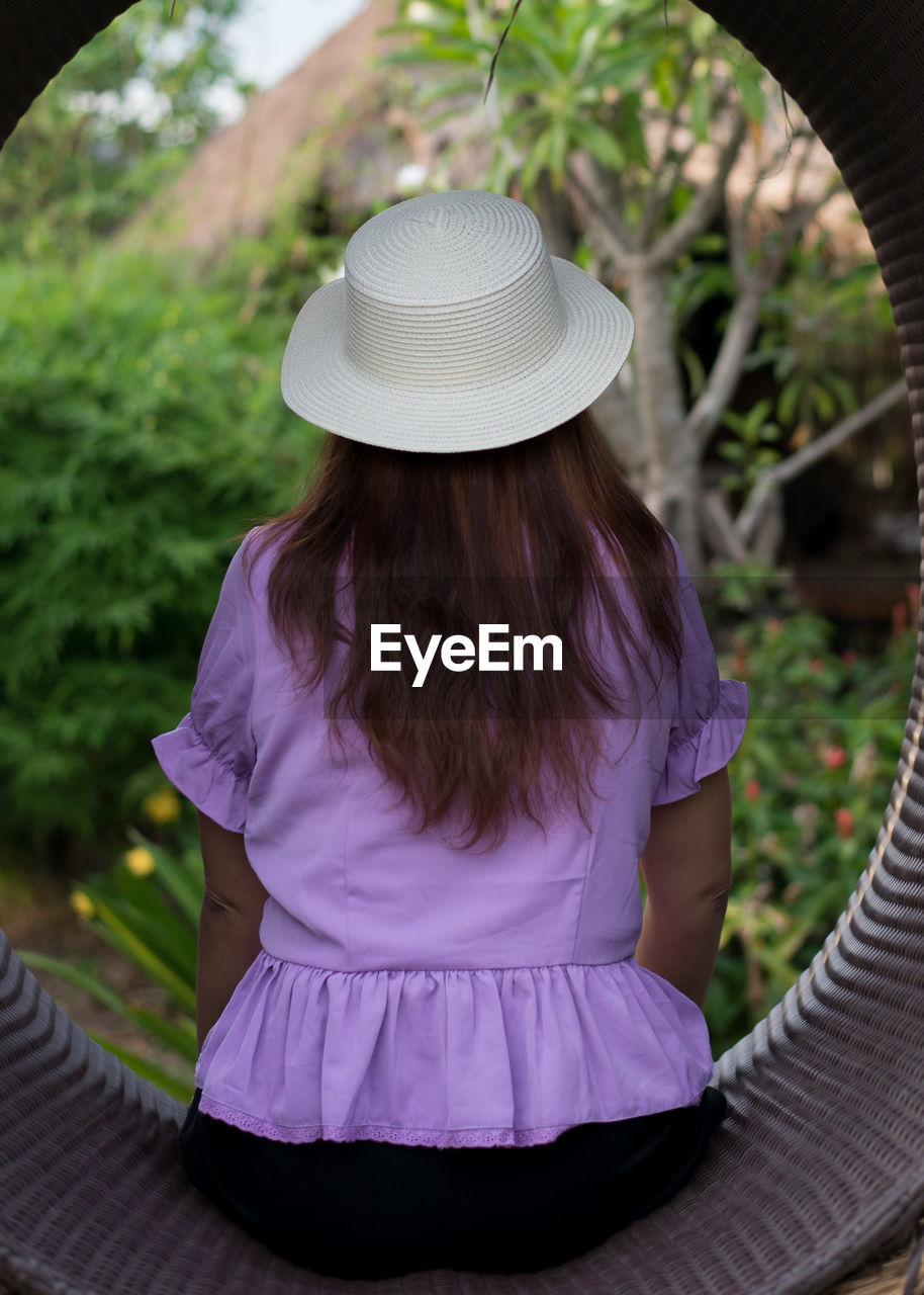 REAR VIEW OF WOMAN WITH HAT SITTING ON PLANT