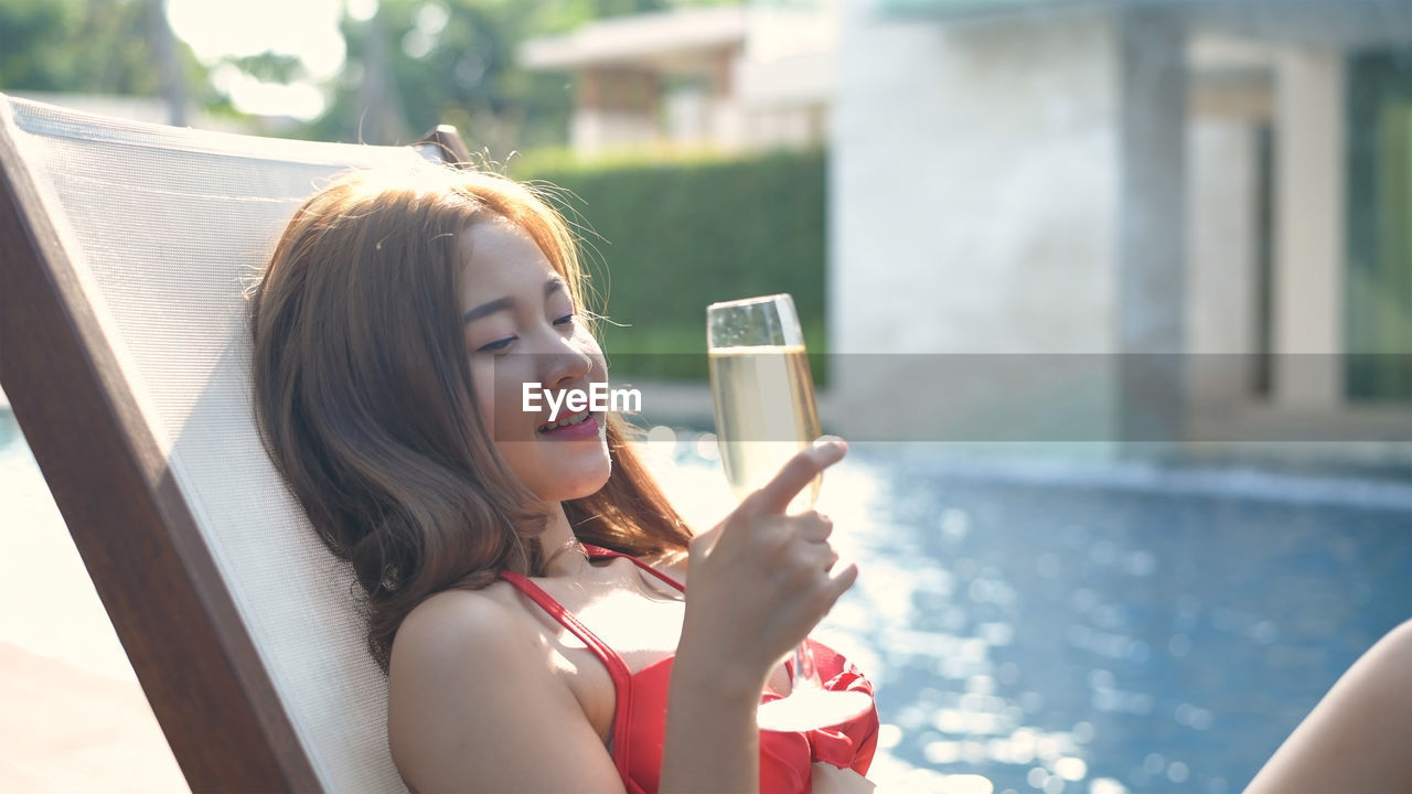 YOUNG WOMAN IN DRINKING GLASS AT SWIMMING POOL AGAINST BLURRED BACKGROUND